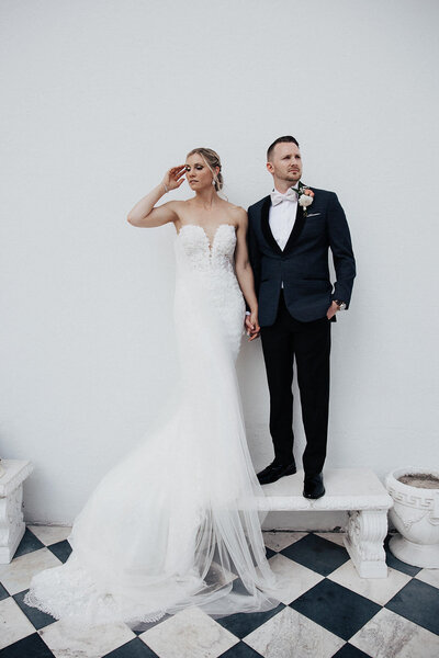 Bride and groom standing on white bench looking in opposite directions. White wall with chess board flooring