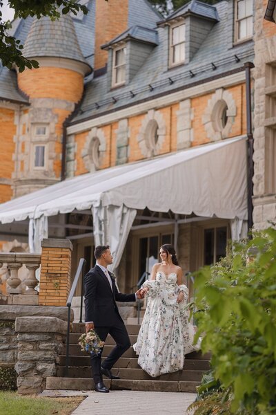 Groom holding his bride's hand as he leads her down the stairs as they walk through the exterior of the Cairwood estate that looks like a castle.