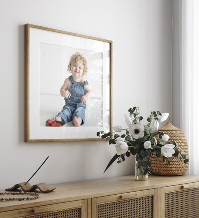 Large framed portrait of a curly-haired toddler in denim overalls, styled on a modern wood console with floral accents.