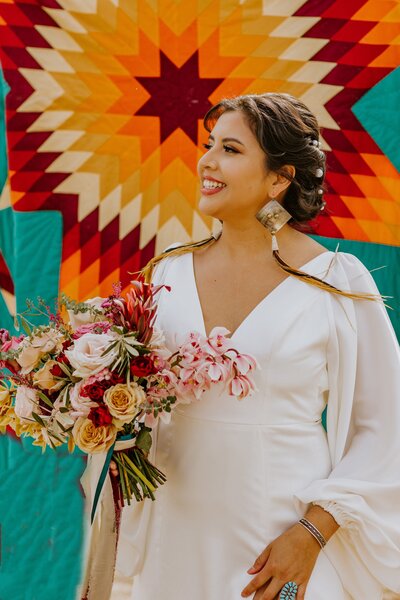 Bride smiles wearing wedding attire with a bouquet and colorful blanket in the background.