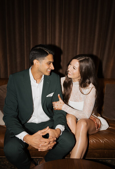 Bride and groom sit and smile at each other in wedding attire.