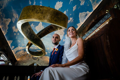 a couple sat on a church pew looking off camera, with the art sculpture in the background at the mount without in bristol