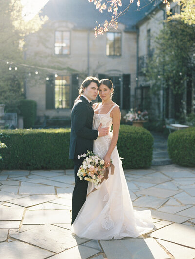 A bride and groom stand closely, facing each other in front of a wooden door with ivy-covered stonework. The groom wears a black tuxedo, and the bride holds a bouquet of flowers. The professional photographer from Greencrest captures every precious moment of this perfect wedding day.