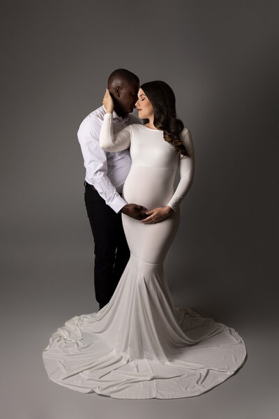 Studio maternity photo shoot - Expectant mom is wearing a long, white maternity gown facing the camera with dad behind. Their heads are touching. Mom and dad both have one hand touching the baby bump, moms other hand is wrapped around dad's neck.