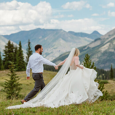 Eloping couple walks hand in hand away from an alpine lake with mountains in the background.