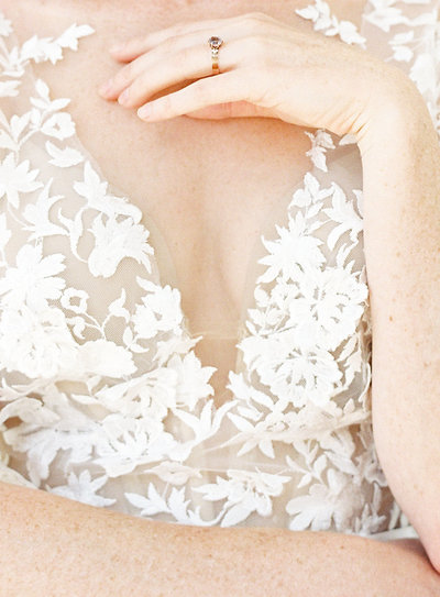 Up close shot of bride's gown from shoulders to hips. One arm is crossing her hips while the other is bent with her left hand relaxing at her collar bones and showing her wedding ring. Gown has 3D printed flowers and leaves on it. Bodice is sheer and 3D print is white. Photographed by wedding photographers in Charleston Amy Mulder Photography