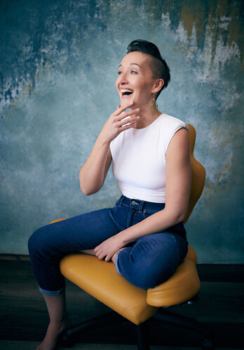 Tarzan, a white woman wearing skinny jeans and a tight white sleeveless shirt with shoulder pads, looks off into the distance laughing, her hand to her chin in a gesture of surprise