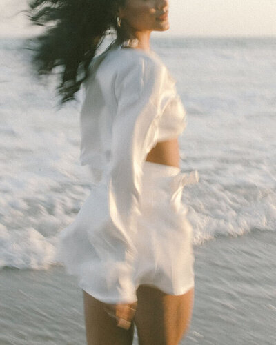 A woman in white, joyfully moving by the beach, embodying the flow of life and spiritual connection.