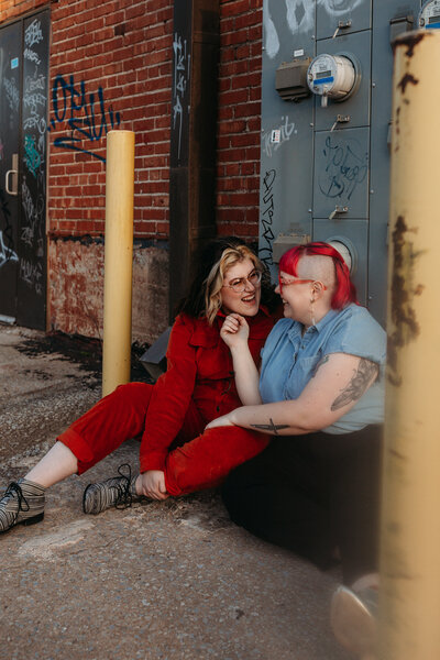 Two people with colorful hair, dressed in casual clothing, share a moment of laughter while sitting against an urban backdrop with graffiti