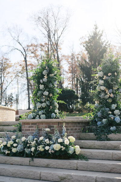 Elegant outdoor wedding ceremony setup featuring lush floral arrangements of white and blue blooms cascading over stone steps, surrounded by trees."