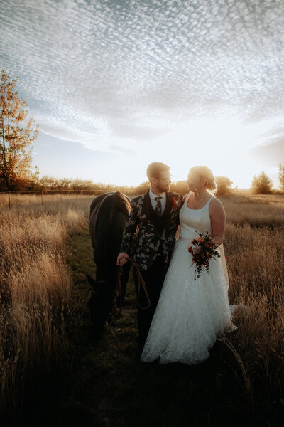 A bride and groom walk toward the camera in a grassy field, leading a horse with them as they look at each other.