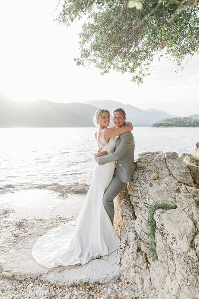 Bride and groom exchanging vows by the water under an olive tree, surrounded by lush white floral arrangements at their intimate destination wedding in Greece