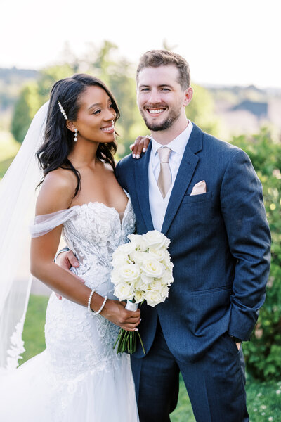 Bride in a lace off-shoulder gown and groom in a navy suit pose together outdoors with soft smiles and a white bouquet, captured during golden hour near Pittsburgh.