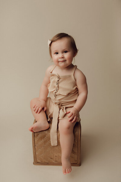 a 1 year old girl wearing a cream outfit sitting on a wicker basket