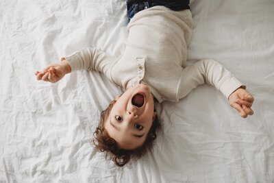 Child lying upside-down on a bed, looking at the camera with an open-mouthed smile during a family photos Pittsburgh session.