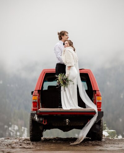 A bride and groom stand on the back of their truck while hugging on their winter mountain wedding day. 