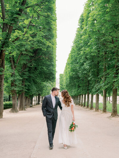 a couple holding hands walking down a row of trees towards the camera at jardin du luxembourg