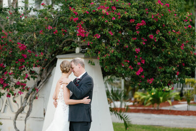 Bride and groom hugging while touching foreheads, standing in front of a large bougainvillea bush