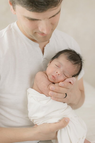 A new father holding newborn baby in his hands in an all white New Jersey studio