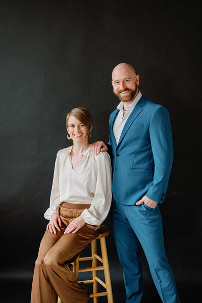 man in a blue suit standing behind a woman sitting on a stool smiling at the camera