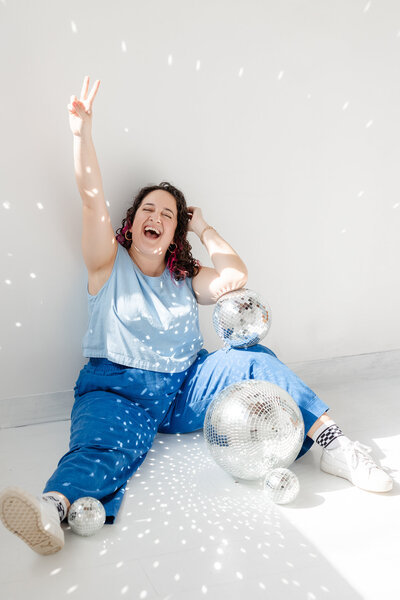 laura sits on the ground, surrounded by disco balls, with a peace sign held in the air