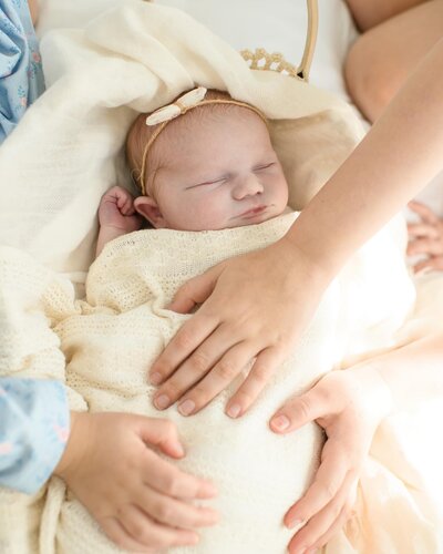 Newborn baby swaddled in a soft blanket sleeps as siblings gently place hands on them. A tender newborn session by Moments in Light Photography.
