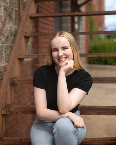 Senior girl with a black shirt and jeans sitting on metal staircase resting her chin on her hand.