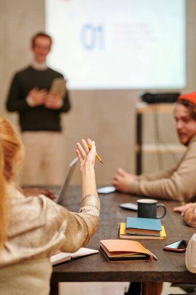 Workshop participant raising hand during corporate training session with presenter in background, books on desk, showing active engagement in learning.