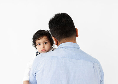 Image of One-year-old baby looking over his dad's shoulder for a photo during a studio cake smash session by Lauren Vanier Photography