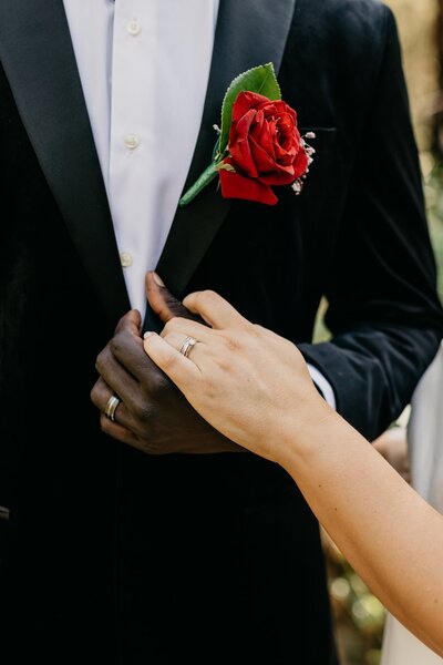 close up of a bride and groom holding hands, showing wedding rings with a red rose in the grooms buttonhole