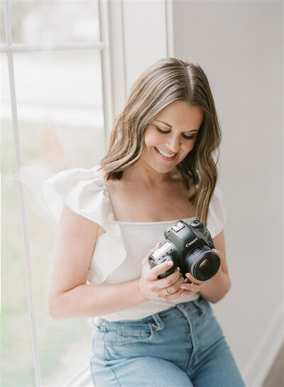 Portrait of a smiling woman holding a professional camera while standing by a window in a bright studio — Kansas City motherhood photographer.