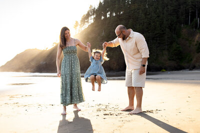 Black and white photo of a family standing barefoot on the beach in front of large coastal rock formations, sharing a quiet moment together.