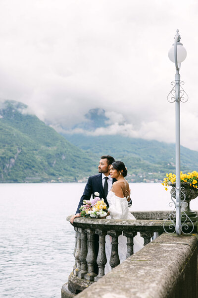 A wedding photo of a couple standing on a stone balcony looking out at lake como from villa aura del lago italy