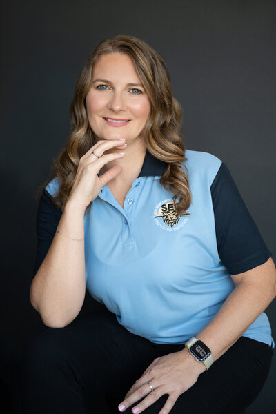 Blond women sitting on a chair wearing a blue polo shirt with her right hand is touching her chin and smiling