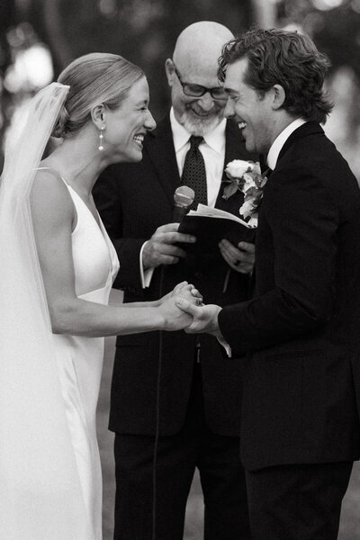 A bride and groom holding hands and smiling as they say their vows