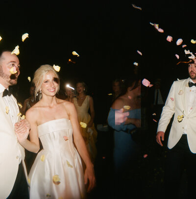 A bride and groom in formal attire walk hand-in-hand through scattered flower petals at night, surrounded by smiling guests.