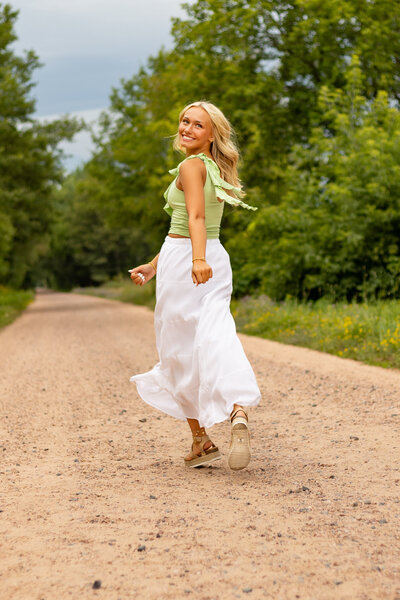 A joyful blonde high school senior twirls on a dirt road, looking back with a radiant smile. She wears a flowing white skirt and a pastel green ruffled top, with the wind catching her outfit for a dreamy, carefree feel. The lush greenery and golden-hour lighting create a soft, natural backdrop, perfect for senior photography inspiration.