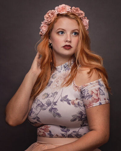 Lady touching hair with right hand and looking left with a crown of flowers over her orange-coloured hair