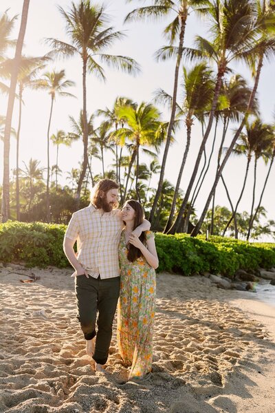 A woman and man hug each other as they walk along the beach with palm trees in the background.