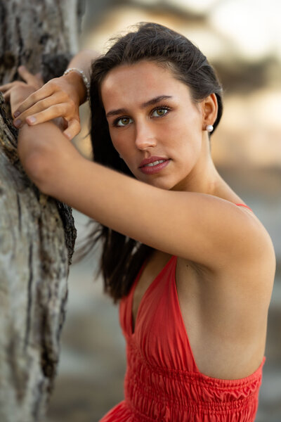 A stunning brunette senior girl poses gracefully against a textured tree trunk, gazing into the camera with captivating green eyes. She wears a bold red sleeveless dress, highlighting her natural beauty and confidence. The warm, golden-hour lighting and softly blurred background create a dreamy, cinematic effect. Perfect for high school senior portrait inspiration, showcasing elegance, depth, and a timeless aesthetic.