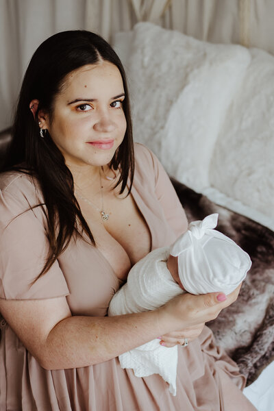 Photo of newborn mom in pink dress sitting on a bed holding newborn baby girl wrapped in white while looking at the camera in a studio in Annapolis Maryland.