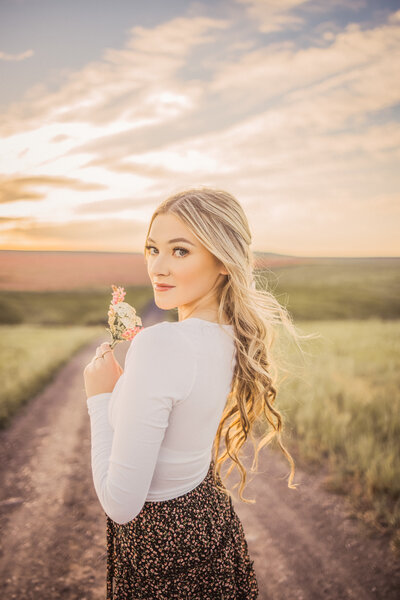 senior girl with long blonde hair holding a small bouquet of wild flowers looking over her shoulder while standing on a dirt road
