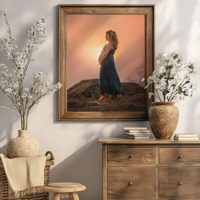 Image of a photo on the wall above a wooden dresser with white flowers. The women in the photo is pregnant wearing a blue dress, holding her belly with the sunset in the background.