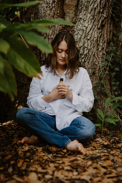 Reiki practitioner wears a white button down and blue jeans sitting on the forest floor holding palo santo to cleanse her energetic field.