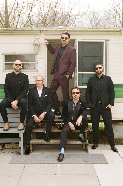 Five men in suits pose on the steps of a trailer captured by wedding film photographer, Jennifer van Son, at Farmhill in Peterborough. One stands while the others sit, all wearing sunglasses, framed by trees in the background.