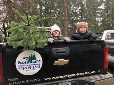 A tree and two young kids are seen in back of a black Ecosystems transfer and recycling pickup truck.