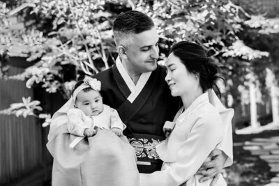 Mother is looking at her youngest son, who is held by his father, lovingly while the oldest boy is looking at the camera with a smile