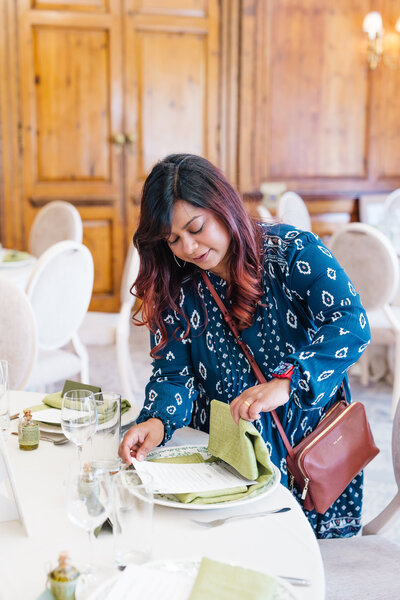 A luxury wedding planner of British-Asian (Indian) heritage, dressed elegantly in a navy blue dress, is making final checks on the wedding breakfast table at Hedsor House in Buckinghamshire. She moves with precision and grace, adjusting table settings, ensuring floral arrangements are perfect, and adding the final touches to create a flawless dining experience. The grand venue’s high ceilings, ornate chandeliers, and large windows fill the room with natural light, enhancing the opulent atmosphere of this beautifully curated celebration.
