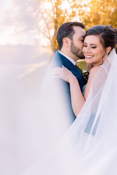 groom kisses his brides temple as her wedding veil swoops towards the camera
