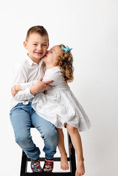 Two siblings kiss and hug on a white backdrop.